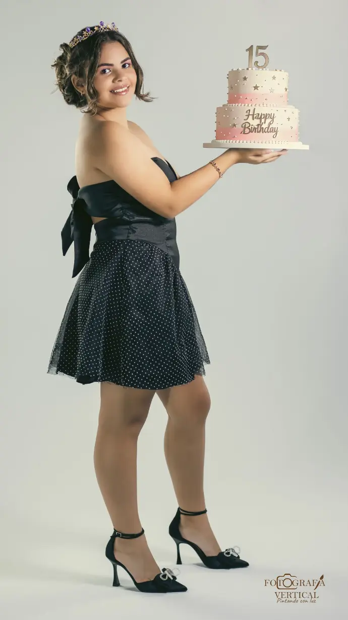 "Retrato de una quinceañera con un elegante vestido negro, sosteniendo una tarta rosa pastel con velas del número 15 y una corona de princesa. La sesión se realiza sobre un fondo claro y suave en tonos pastel."