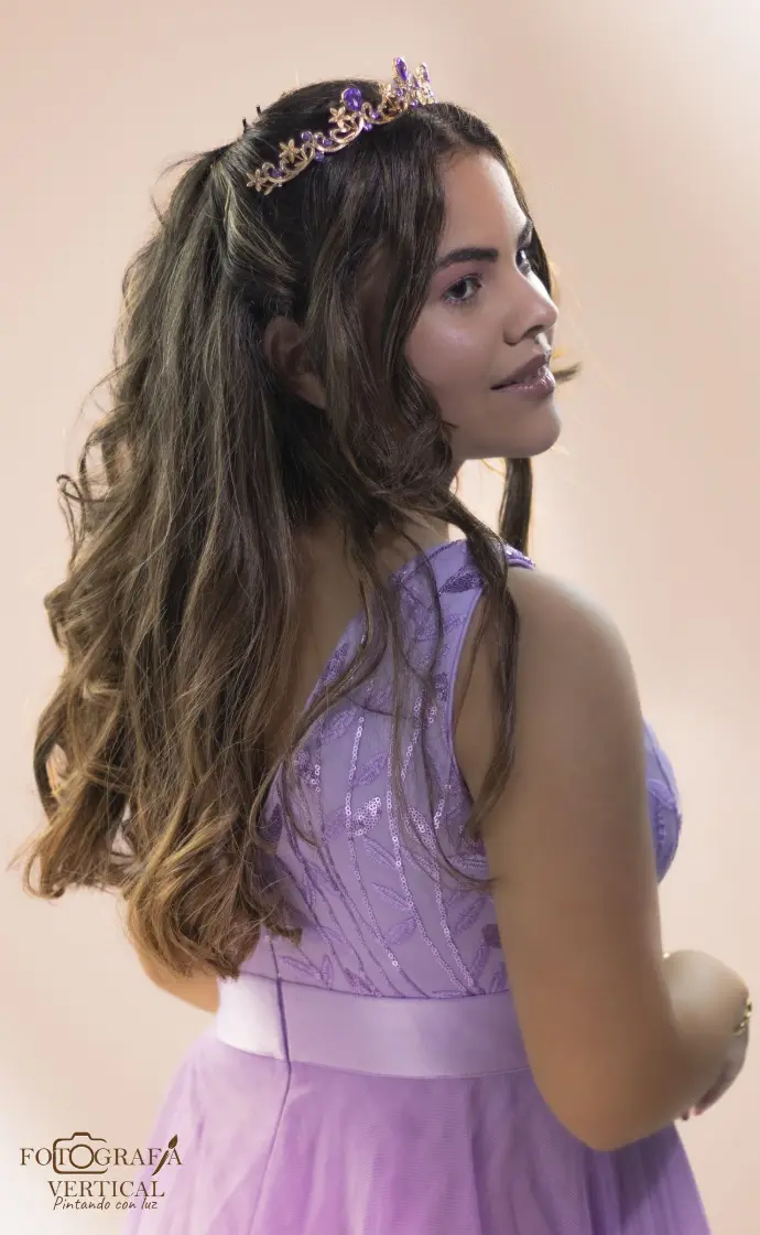 Retrato de cerca de la joven de espalda, mostrando el peinado de ondas sueltas y la espalda de un vestido lila de encaje y tul, destacando una tiara con gemas moradas, con un fondo rosado cálido y suave.