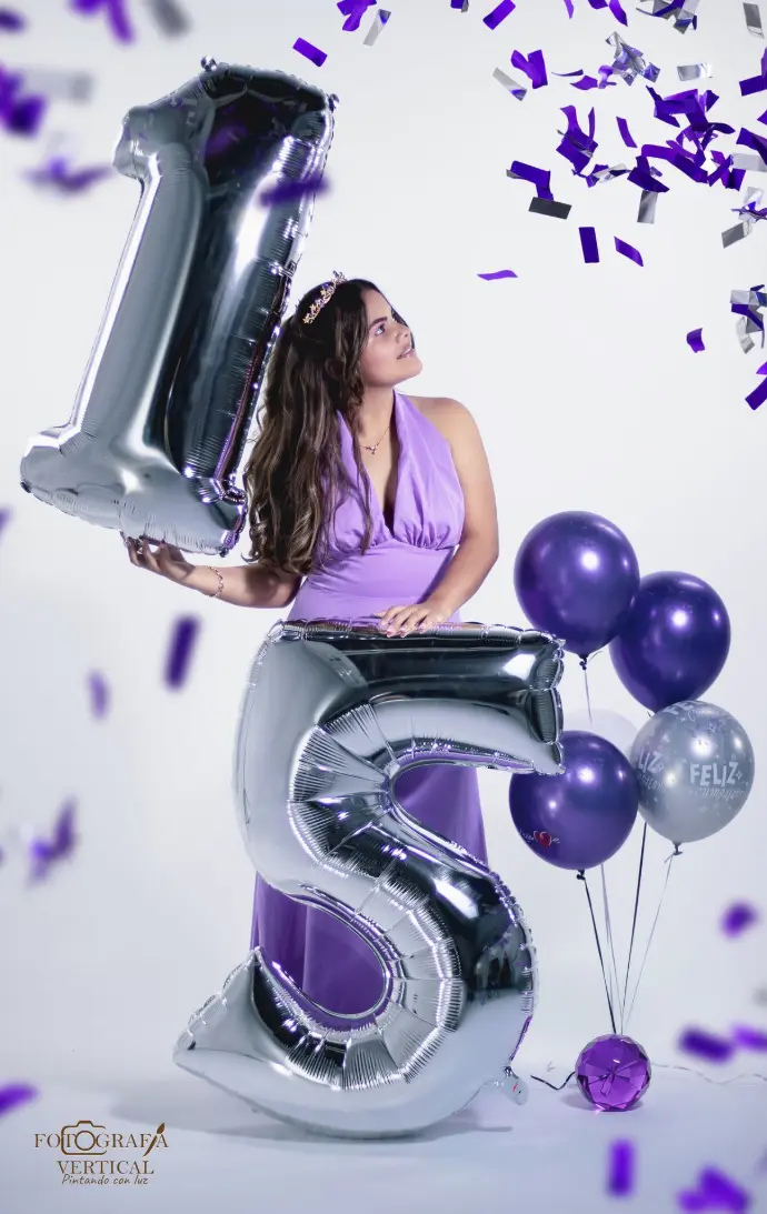 "Quinceañera radiante mira cómo caen lentamente purpurina violeta y plateada, en una sesión de fotos con fondo blanco impecable y globos conmemorativos. Algunas partículas de purpurina aparecen desenfocadas, creando un efecto mágico y onírico."
